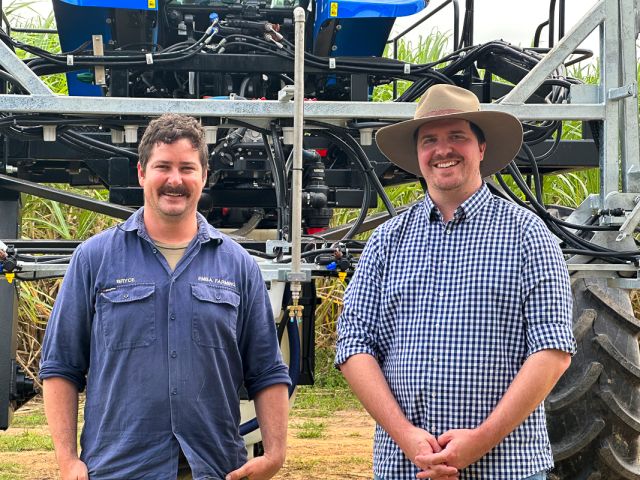 Two men pose in front of a green tractor, smiling and wearing work clothes in an outdoor setting. 