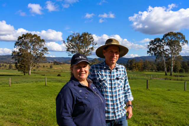 A man and woman stand together in a green field under a clear blue sky, smiling and enjoying the outdoors. 
